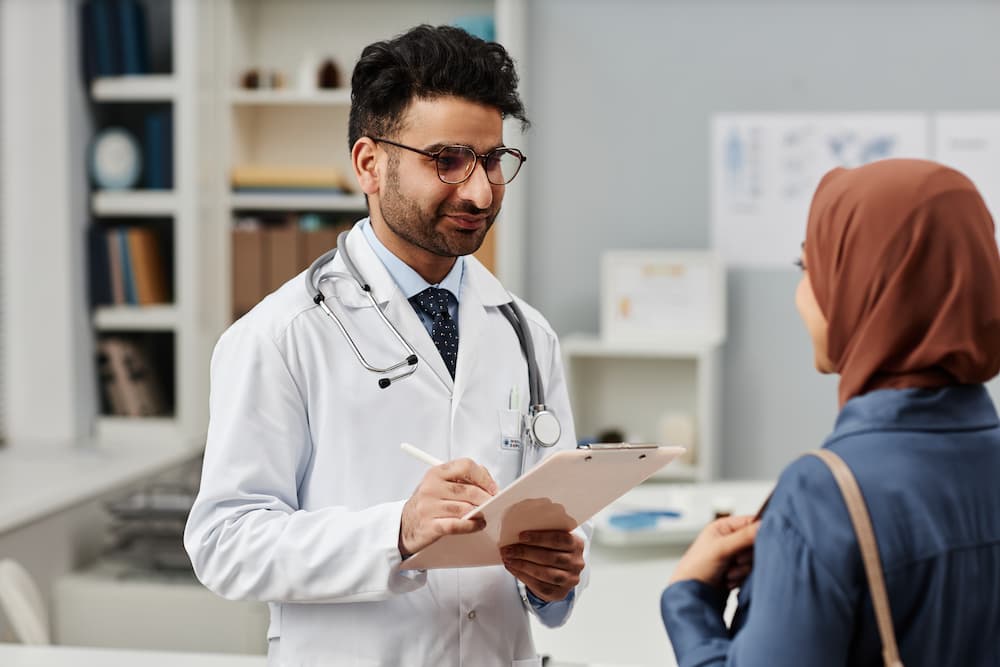 Male doctor with a clipboard talking to a female patient in a headscarf in a medical office.